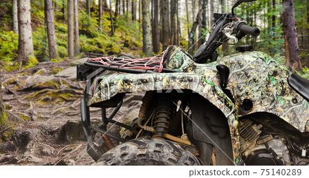 Photo of a camouflaged offroad hunting atv vehicle standing in forest profile close-up view. 75140289