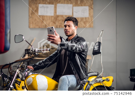 A stylish guy on a yellow cool motorcycle takes a selfie in a garage or car repair shop A stylish guy on a yellow cool motorcycle takes a selfie in a garage or car repair shop 75142946