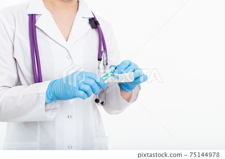 A young nurse in a mask opens a syringe on a white background in a white coat, to make a vaccination A young nurse in a mask opens a syringe on a white background in a white coat, to make a vaccination 75144978