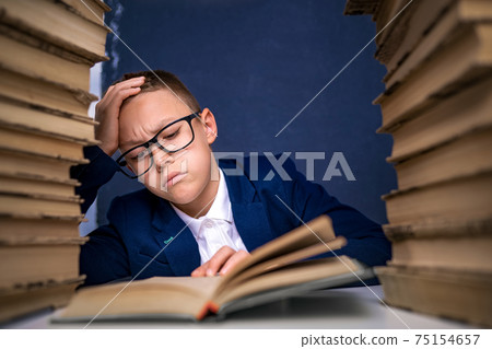 Smart boy in glasses sitting between two piles of books and read book thoughtfully 75154657