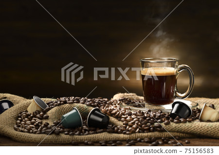 Cup glass of coffee with smoke, coffee beans and coffee capsules on burlap sack on wooden background Cup glass of coffee with smoke, coffee beans and coffee capsules on burlap sack on wooden background 75156583