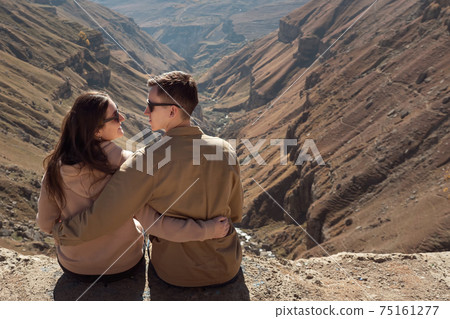 Romantic couple in love hugs sitting on mountain cliff edge Romantic couple in love hugs sitting on mountain cliff edge 75161277