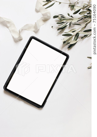 Electronic device mock-up. Closeup of tablet with empty screen. Olive tree branches and silk ribbon isolated on white table background. Flat lay, top view. Wedding design. Home office. Vertical. Electronic device mock-up. Closeup of tablet with empty screen. Olive tree branches and silk ribbon isolated on white table background. Flat lay, top view. Wedding design. Home office. Vertical. 75164090