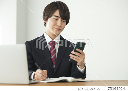 一個穿著西裝,一邊看著智能手機的屏幕做筆記的年輕人 一個穿著西裝,一邊看著智能手機的屏幕做筆記的年輕人 75165924