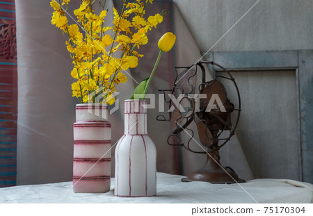 Bouquet of yellow flowers in Two handmade ceramic vase and Vintage fan on white textured table cloth with old cement wall. 75170304