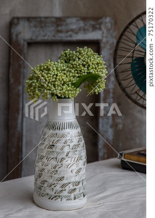 Bouquet of green flowers in White handmade ceramic vase bottle shape and Vintage fan on white textured table cloth with old cement wall. 75170322