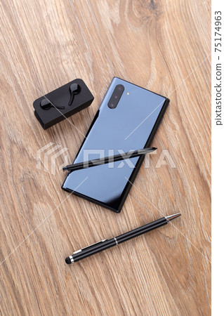 Smartphone with three cameras, stylus, pen and headphones on a wooden table close-up. 75174963