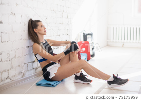 Girl in the gym is wrapping up boxing bandages on her hands before training a blow on a boxing bag Girl in the gym is wrapping up boxing bandages on her hands before training a blow on a boxing bag 75175959
