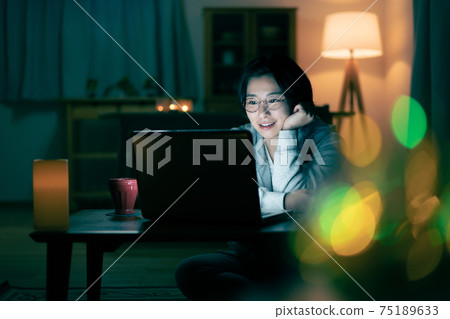 A smiling young woman looking at a computer in the living room at night A smiling young woman looking at a computer in the living room at night 75189633