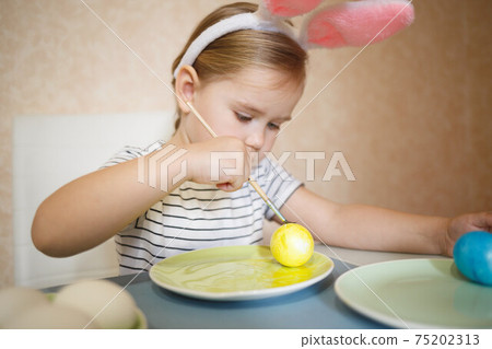 little cute blue-eyed girl in bunny ears paints Easter eggs 75202313