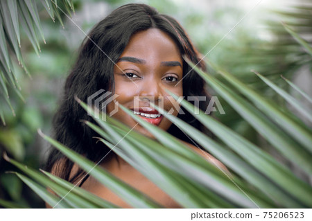 Closeup of young charming African woman with healthy skin and natural nude makeup, posing among palm leaves of green plants in hothouse. Skin care and wellness concept. Closeup of young charming African woman with healthy skin and natural nude makeup, posing among palm leaves of green plants in hothouse. Skin care and wellness concept. 75206523