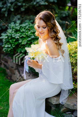 A beautiful bride with long curls sits on a park bench among grass and trees. A beautiful bride with long curls sits on a park bench among grass and trees. 75206761