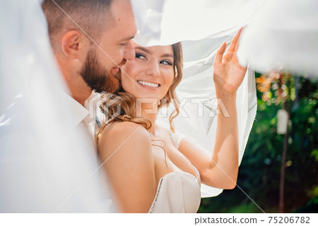 under the veil Happy and beautiful bride and groom gently cuddle in the park 75206782