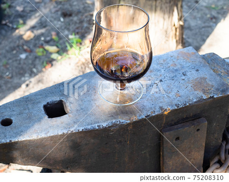 glass of brandy on iron anvil in rural smithy 75208310