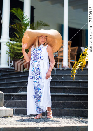 the woman in the big hat is smiling. a beautiful girl in a big hat and white dress smiles outside an old colonial building on the island of Mauritius 75209124