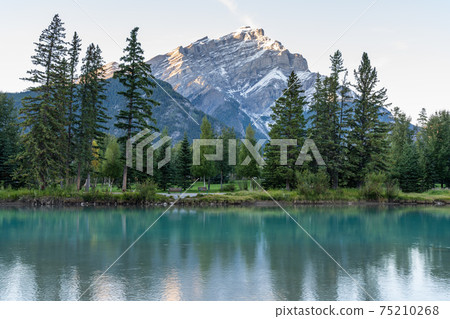 Banff National Park beautiful scenery. Cascade Mountain and pine trees reflected on turquoise color Bow River in summer time. Town of Banff, Canadian Rockies, Alberta, Canada. Banff National Park beautiful scenery. Cascade Mountain and pine trees reflected on turquoise color Bow River in summer time. Town of Banff, Canadian Rockies, Alberta, Canada. 75210268