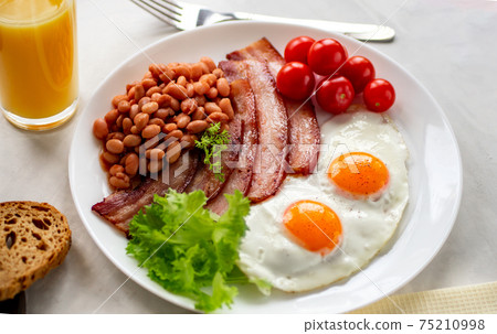 Hearty breakfast with scrambled eggs, bacon, beans, cherry tomatoes, homemade bread, orange juice on a concrete countertop. English breakfast concept. Hearty breakfast with scrambled eggs, bacon, beans, cherry tomatoes, homemade bread, orange juice on a concrete countertop. English breakfast concept. 75210998