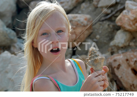Young blonde girl surprised and holding a seashell Young blonde girl surprised and holding a seashell 75217749