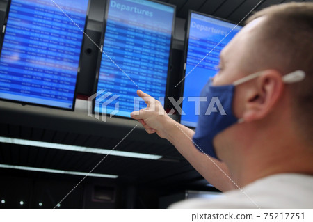 Young man protect his health with a medical mask an airport time table terminal background Young man protect his health with a medical mask an airport time table terminal background 75217751