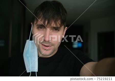Photo of a guy with a thick medical mask on a dark, blurry background. Long bangs, tired smile, grin. Selfie Photo of a guy with a thick medical mask on a dark, blurry background. Long bangs, tired smile, grin. Selfie 75219211