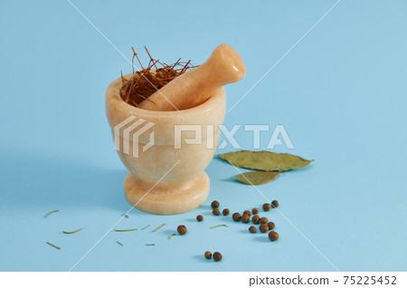 Beige marble mortar with pestle for grinding ingredients near peppercorns and bay leaf lying on a blue background with copy space. 75225452