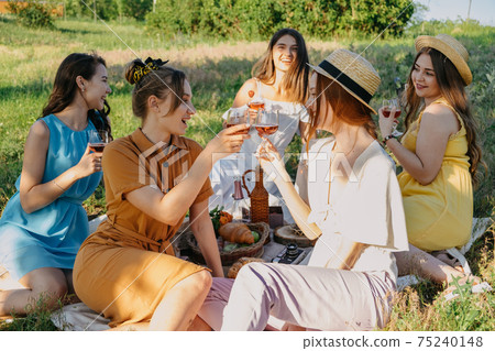 Friends having picnic in the countryside. Group of young women sitting on blanket in park near trees, at sunset on spring summer day. Five girlfriends eating and drinking red wine on outdoor party Friends having picnic in the countryside. Group of young women sitting on blanket in park near trees, at sunset on spring summer day. Five girlfriends eating and drinking red wine on outdoor party 75240148