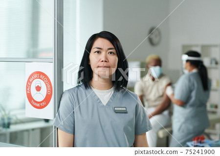 Young female clinician in uniform standing in front of camera against colleague vaccinating patient 75241900