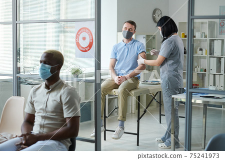 Brunette nurse in uniform and protective mask making injection to young man 75241973