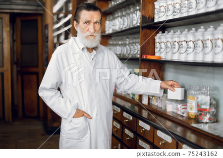 People, healthcare, pharmacy concept. Indoor portrait of pleasant professional mature bearded man pharmacist, wearing white coat, posing in beautiful old pharmacy interior People, healthcare, pharmacy concept. Indoor portrait of pleasant professional mature bearded man pharmacist, wearing white coat, posing in beautiful old pharmacy interior 75243162