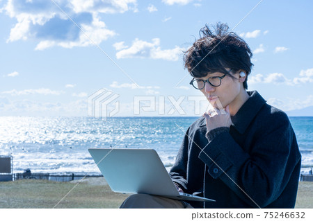 A man using a laptop while wearing earphones at the beach 75246632