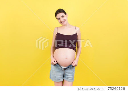 Happy pregnant woman in unzipped jeans showing her naked abdomen at colorful background with copy space 75260126