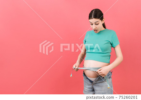 Cropped image of expecting mother in unzipped jeans measuring her pregnant belly with a tape measure at colorful background. Centimeter measurement Cropped image of expecting mother in unzipped jeans measuring her pregnant belly with a tape measure at colorful background. Centimeter measurement 75260201