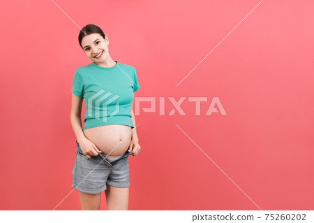 Happy pregnant woman in unzipped jeans showing her naked belly at colorful background with copy space. Baby expecting concept Happy pregnant woman in unzipped jeans showing her naked belly at colorful background with copy space. Baby expecting concept 75260202