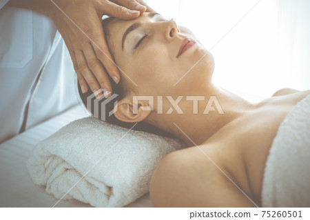 Beautiful brunette woman enjoying facial massage with closed eyes in sunny spa center. Relaxing treatment and cosmetic medicine concepts 75260501