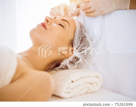 Beautiful woman receiving beauty injections with closed eyes in sunny medical center. Beautician doctor hands doing beauty procedure to female face with syringe. Cosmetic medicine and surgery concept 75261099