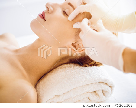 Beautiful woman receiving beauty injections with closed eyes in sunny medical center. Beautician doctor hands doing beauty procedure to female face with syringe. Cosmetic medicine and surgery concept Beautiful woman receiving beauty injections with closed eyes in sunny medical center. Beautician doctor hands doing beauty procedure to female face with syringe. Cosmetic medicine and surgery concept 75261102