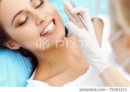 Young female patient visiting dentist office.Beautiful woman with healthy straight white teeth sitting at dental chair with open mouth during oral checkup while doctor working at teeth Young female patient visiting dentist office.Beautiful woman with healthy straight white teeth sitting at dental chair with open mouth during oral checkup while doctor working at teeth 75261211