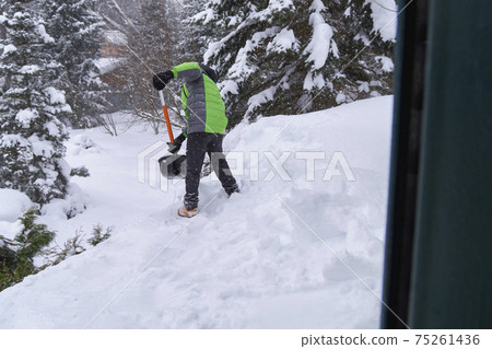 Snow removal on the roof house. Boy in green jacket with shovel throws snow off the roof. 75261436
