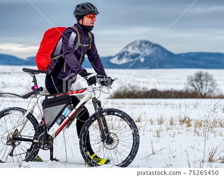Cyclist in black sport suit pushing the mountain Bike on the Snowy Trail. 75265450
