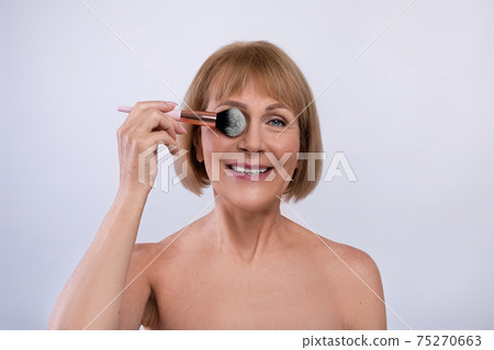 Natural makeup for aging skin. Portrait of lovely mature lady holding cosmetic brush near her eye on light background Natural makeup for aging skin. Portrait of lovely mature lady holding cosmetic brush near her eye on light background 75270663