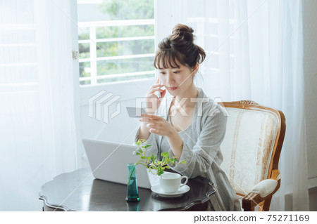 一個穿著睡衣的年輕女子,看著她的電腦前的卡片時,在她的智能手機上進行查詢 一個穿著睡衣的年輕女子,看著她的電腦前的卡片時,在她的智能手機上進行查詢 75271169