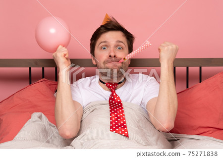 man celebrating his birthday, blowing party wistle and rejoicing. He stay at bed at home alone. man celebrating his birthday, blowing party wistle and rejoicing. He stay at bed at home alone. 75271838