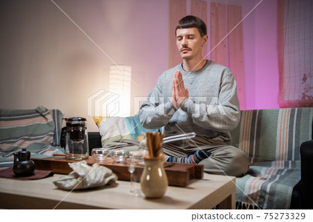 A man meditates to achieve harmony with the world around him, before starting a traditional oriental tea party at the tea table and bamboo tray. A man meditates to achieve harmony with the world around him, before starting a traditional oriental tea party at the tea table and bamboo tray. 75273329
