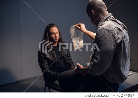 In the interrogation room, an officer of the drug control service presents evidence of white cocaine powder in a package to the criminal. 75274084