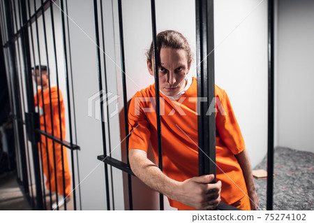 Harsh wild prisoner criminal maniac with tattoos grabs the bars. A close-up portrait in a prison cell. 75274102