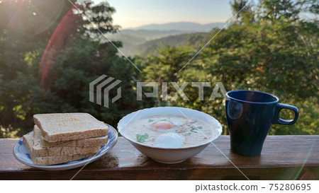 rice porridge with pork and boiled egg with coriander leaf on white bowl serving with black mug of home made coffee and grilled Whole wheat bread in the middle of forest mountain in Summer season, adv 75280695
