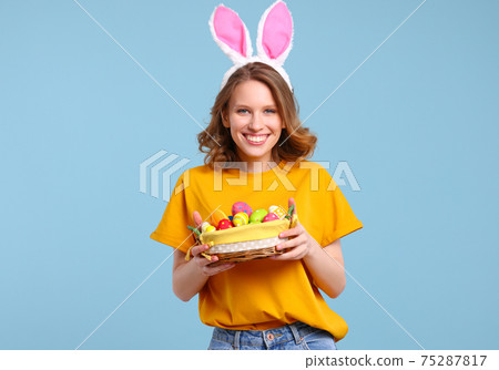 Positive young woman with bunny ears smiling and holding Easter basket in hands Positive young woman with bunny ears smiling and holding Easter basket in hands 75287817