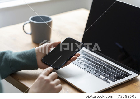 Hands of a man holding a smartphone in front of a laptop 75288503