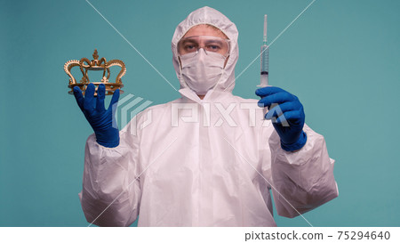 A male doctor in protective overalls and a mask holds an syringe with a vaccine and crown in hand 75294640