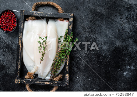 Raw white Squid or Calamari in a wooden tray with herbs. Black background. Top view. Copy space 75298847